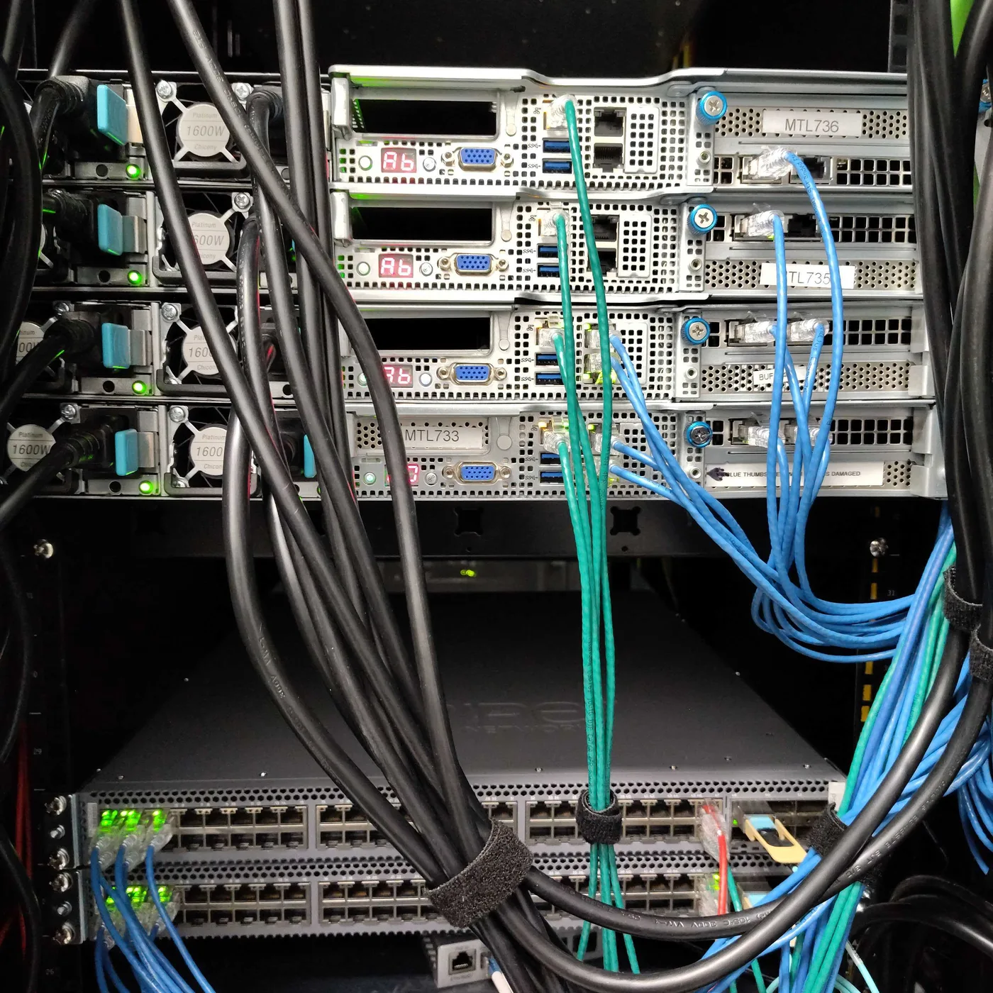 Photograph of an installed cloud rack, rear view: four 1U/2U servers at the top with cable management trays between them, a Juniper QFX5100 switch below, and dense blue and turquoise cabling running between them.